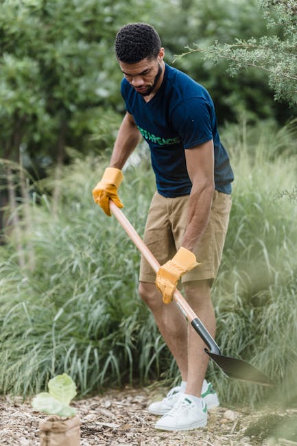 A young man with dark hair and a beard, dressed in a navy blue t-shirt and beige shorts, is engaged in outdoor garden maintenance. He is wearing bright yellow gardening gloves and holding a long-handl
